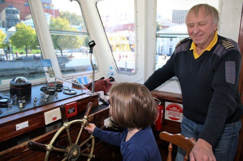 Een persoon aan het roer, een andere persoon ernaast, en een uitzicht op het water en gebouwen door de ramen van het schip. 4
