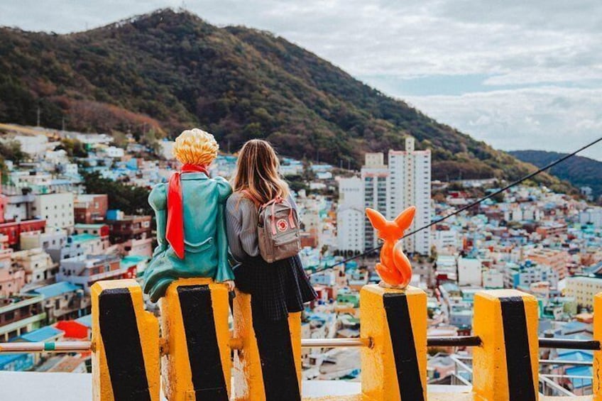 Dos personas sentadas en un saliente, con vistas a una ciudad con edificios coloridos y una colina al fondo. 2