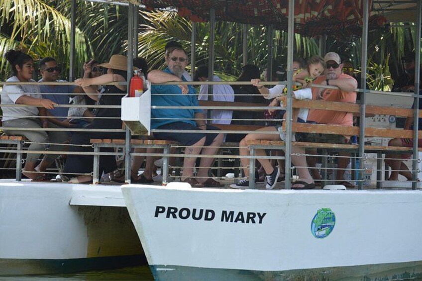 Un bote con gente sentada en la cubierta, una barandilla y el nombre 'PROUD MARY' en el costado. 2