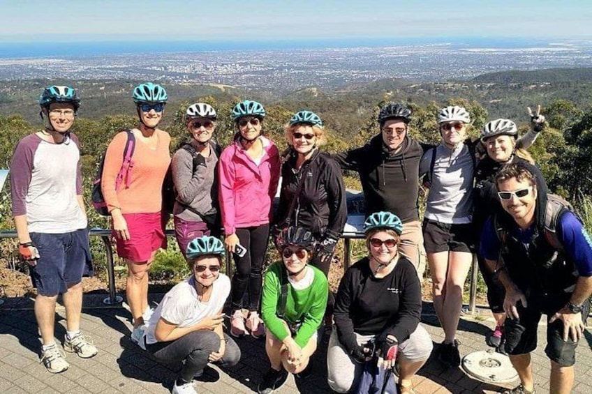 A group of people wearing helmets and cycling attire, posing on a ledge with a cityscape in the background. 1