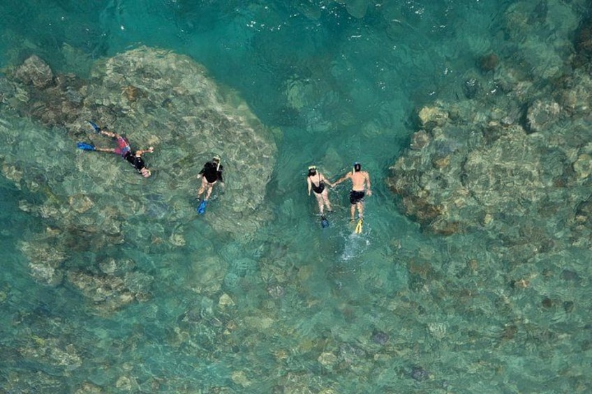Tre personer snorkler i krystalklart turkis vand med synlige undervandssten. 1