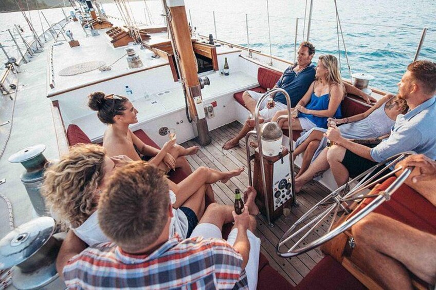 Un groupe de personnes se trouve sur un bateau doté d’un pont en bois, avec une petite table sur laquelle sont posées des boissons, et une personne tient une bouteille. 2