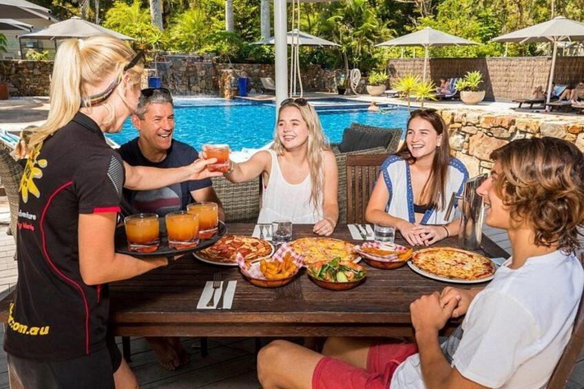 A group of people dining outdoors by a pool with a waiter serving drinks and food. 2