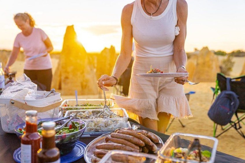 A picnic setup with food and drinks on a table, a person serving food, and a sunset in the background. 3