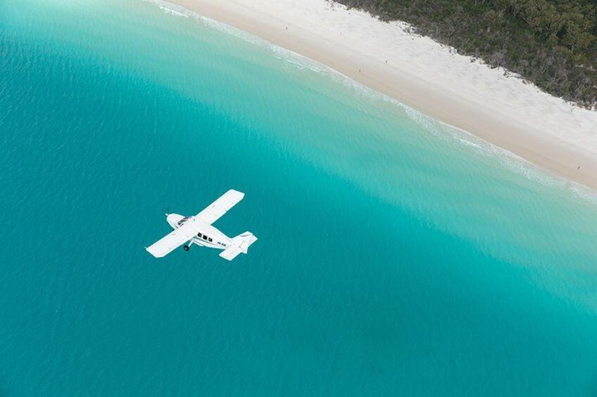 Un pequeño avión blanco vuela sobre un mar turquesa con una playa de arena blanca y una densa vegetación verde a la derecha. 5