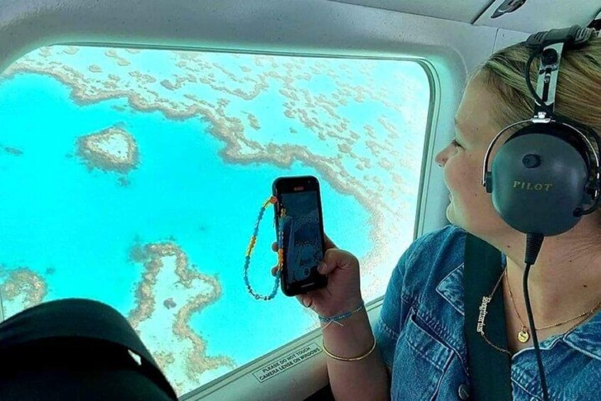 Una persona sosteniendo un teléfono inteligente, usando una chaqueta vaquera y auriculares, con vista a aguas turquesas y arrecifes de coral a través de una ventana de avión. 1