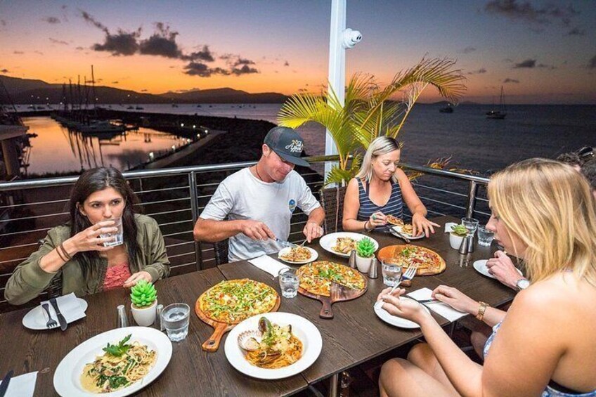 A group of people dining at a table with a view of the ocean and a sunset. 1