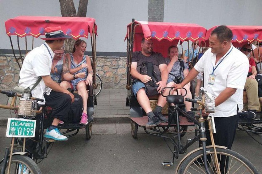 Två rickshaw-cyklar med passagerare, en person som står framför, och en stenmur i bakgrunden. 2