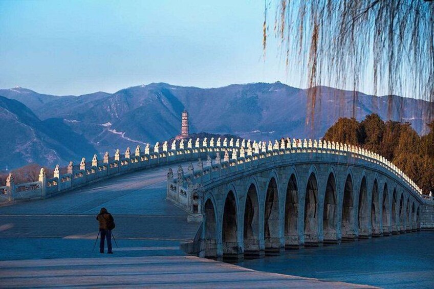 A stone bridge with arches, a person standing on the bridge, and mountains in the background. 2