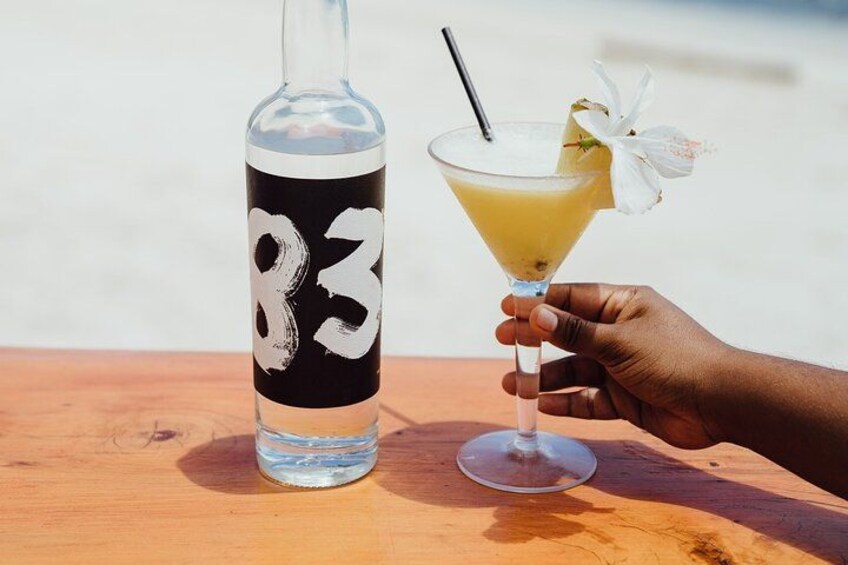 Une main tenant un verre à cocktail avec une paille et une fleur en guise de décoration, à côté d’une bouteille dont l’étiquette est noire et qui porte le numéro 83. 1