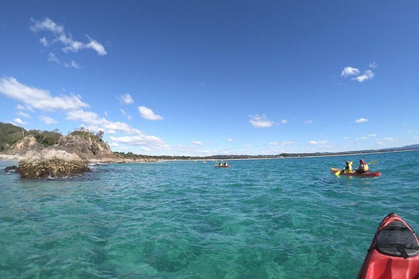 A kayaker in a red kayak, another in a yellow kayak, and a third in a white kayak on a clear blue sea with a rocky outcrop on the left. 4