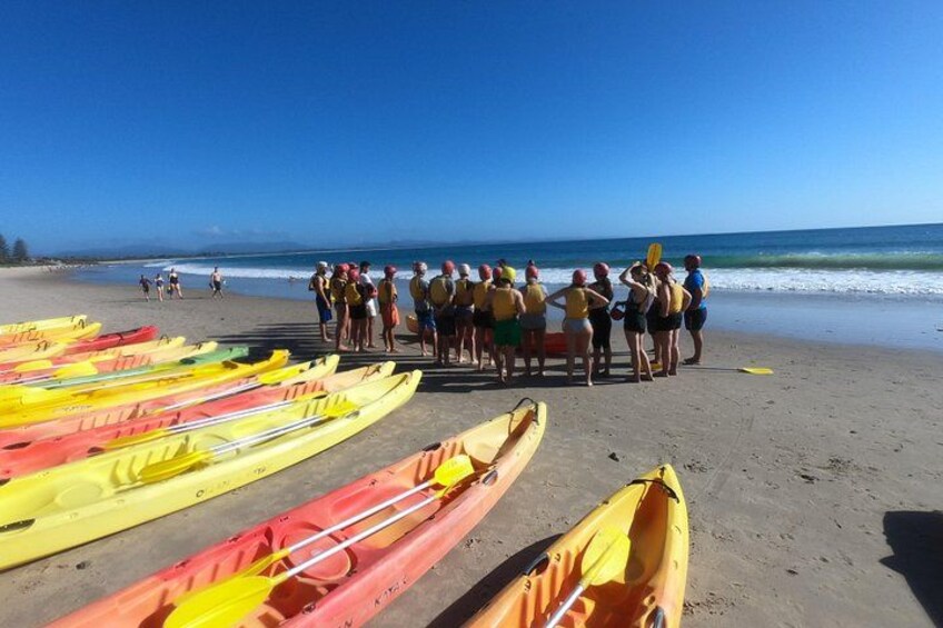 Une plage où des personnes sont rassemblées en groupe, des kayaks alignés sur le sable, et l’océan en arrière-plan. 4