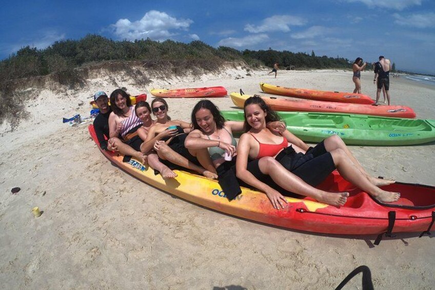 A group of people sitting on colorful kayaks on a sandy beach with a forested hill in the background. 5