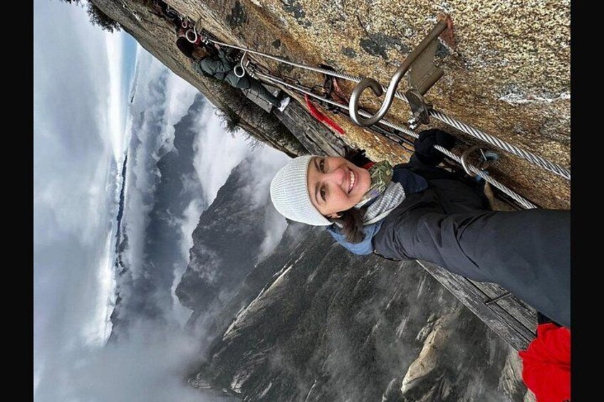 A person standing on a wooden platform with a rope ladder, overlooking a mountainous landscape with clouds. 1