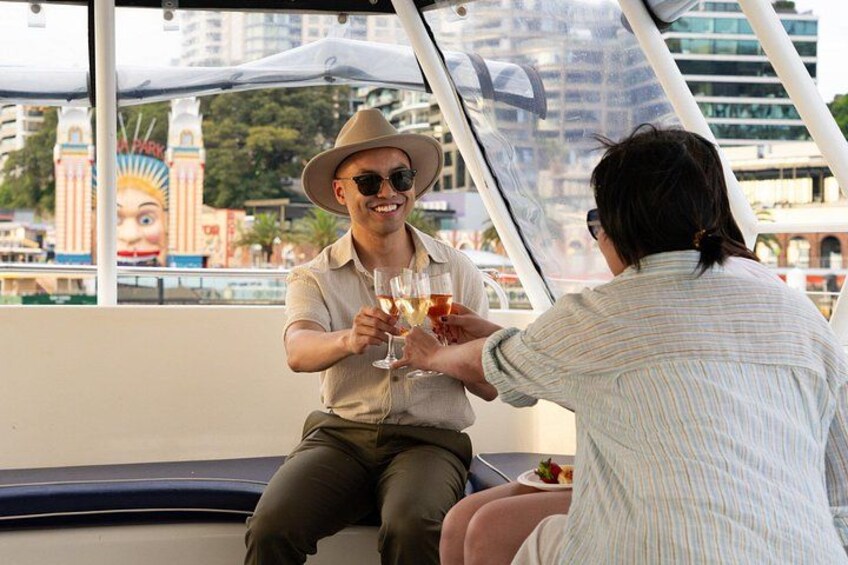 Twee personen proosten met drankjes op een boot, met een stadsgezicht op de achtergrond. 2