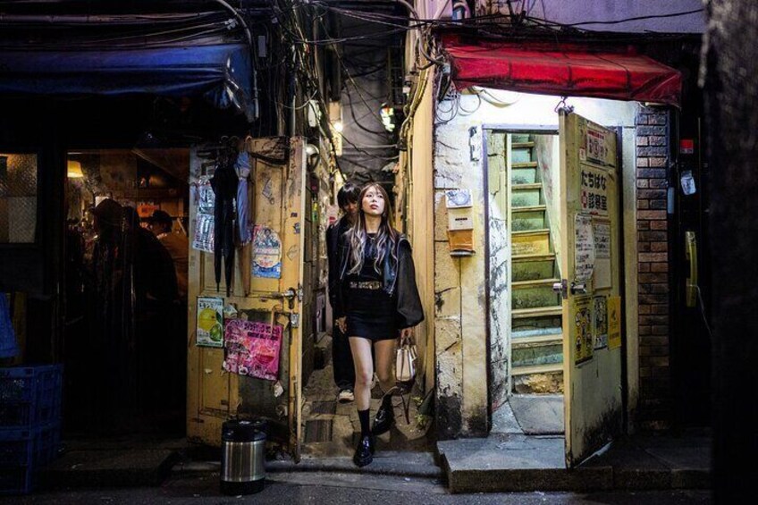 A narrow alleyway with a person walking, a trash can, and a staircase leading to a door with Japanese text. 5