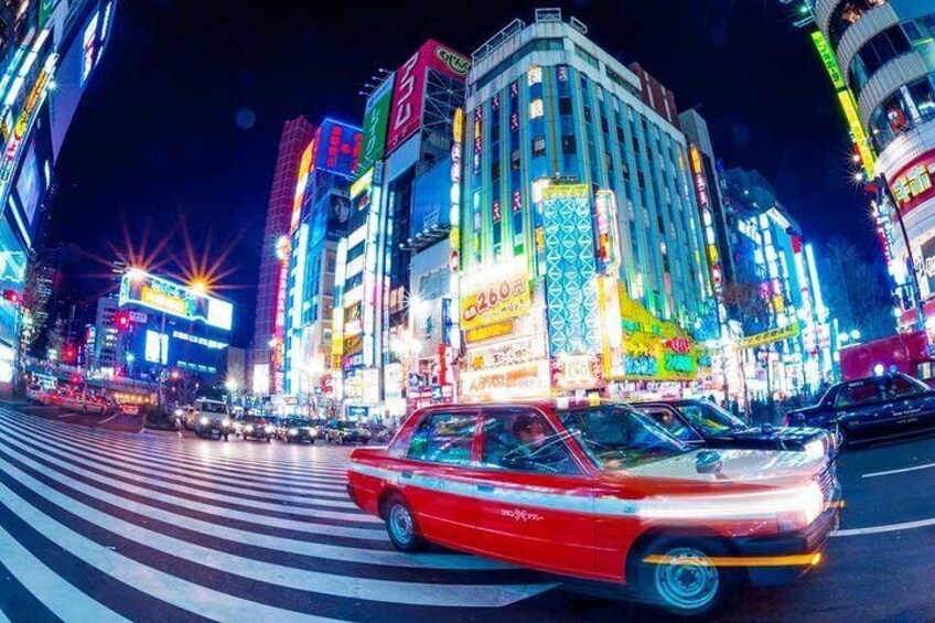 A brightly lit urban street at night with a red taxi in motion, neon signs, and illuminated buildings. 2