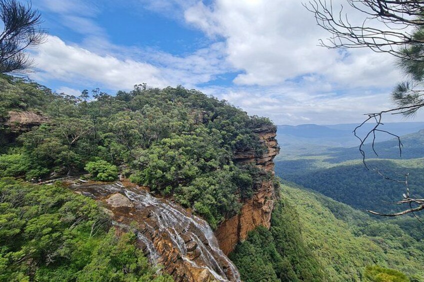 A cliff with a waterfall, dense green forest, and a distant mountain range under a partly cloudy sky. 2