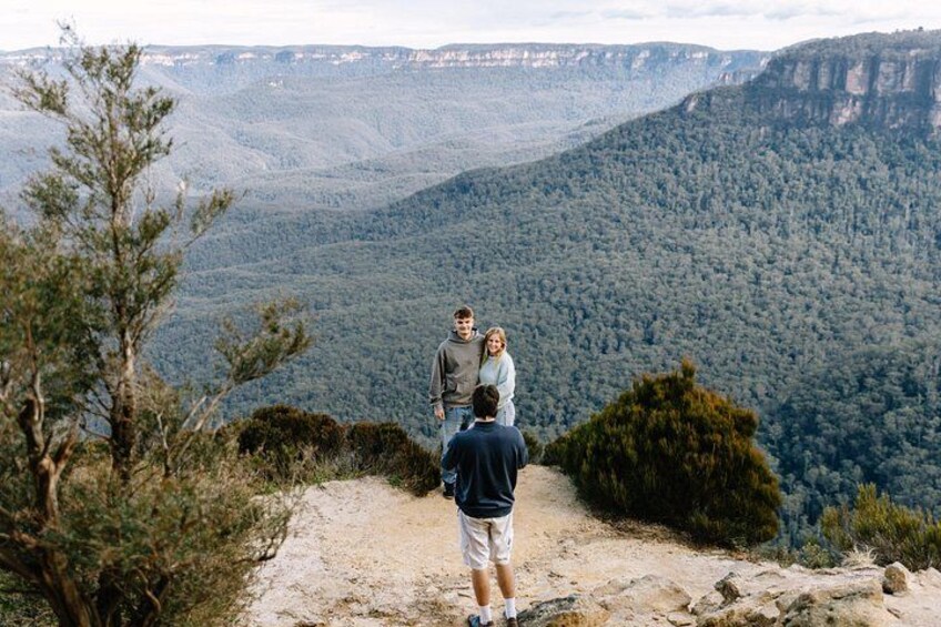 A group of people standing on a dirt path overlooking a forested valley with a mountain range in the background. 1