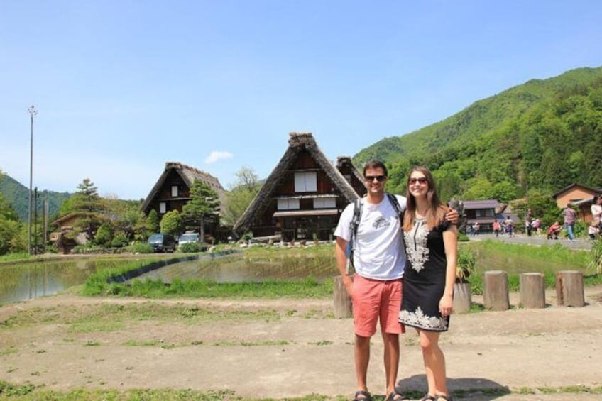 Two people standing in front of traditional buildings with thatched roofs, a body of water, and a hilly landscape in the background. 1