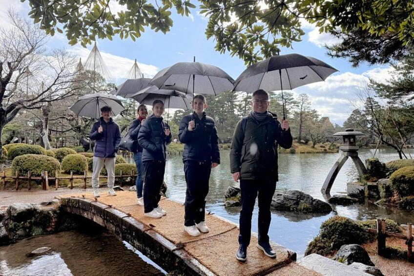 Een groep mensen staat bij een vijver met parasols, omringd door bomen en een stenen brug. 2
