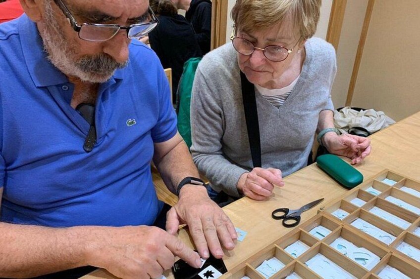 Twee mensen werken aan een houten puzzel, met een schaar en een groene tas in de buurt. 3