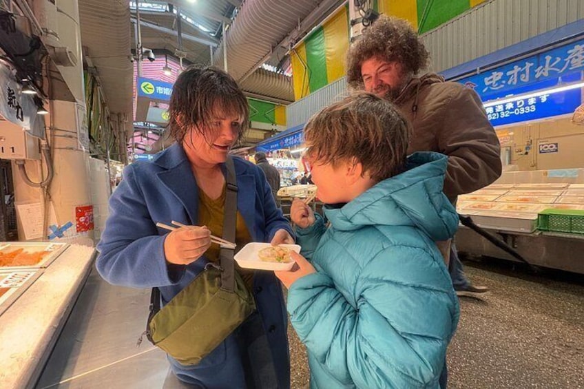 Una persona come sushi en un mercado con varios puestos de comida y letreros al fondo. 2