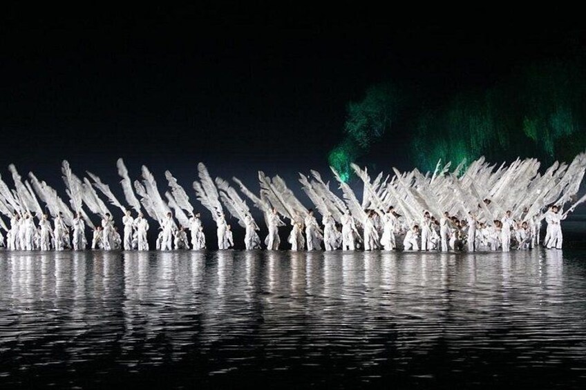 A nighttime outdoor performance with a group of people in white costumes, illuminated by stage lights, standing in formation on a reflective surface. 3