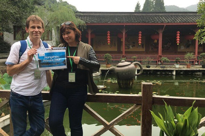 Two people holding a sign with 'Hangzhou Private Tour' written on it, standing by a traditional Chinese building with a pond and a teapot. 2