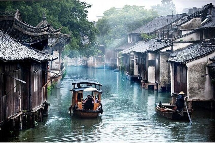 A narrow waterway flanked by traditional wooden buildings with tiled roofs, a boat with passengers, and a person rowing a smaller boat. 1