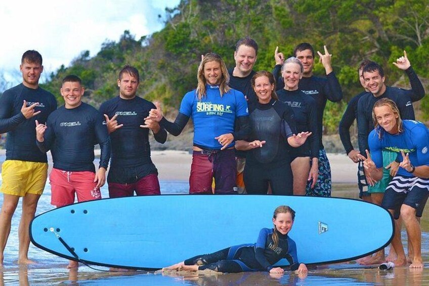 A group of people in wetsuits posing with a surfboard on a beach. 3