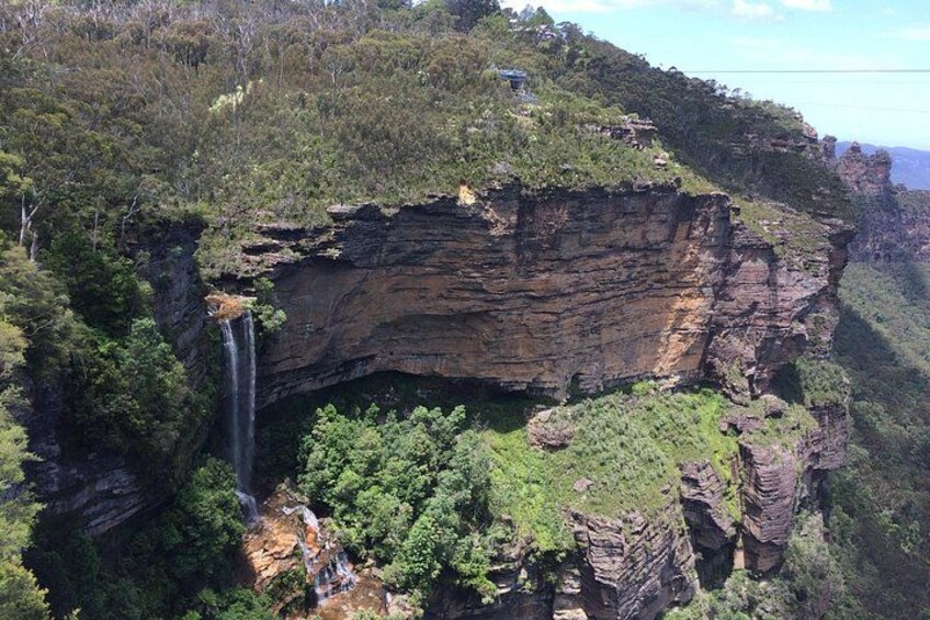 A cliff with a waterfall, dense greenery, and a small building on top. 2