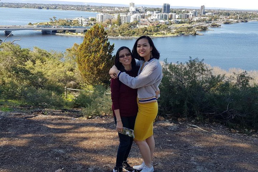 Two people embracing on a dirt path with a river and city skyline in the background. 2