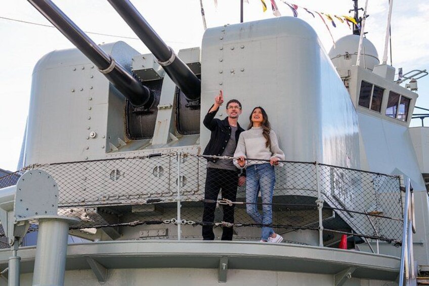 Un couple se tient sur le pont d’un navire, à proximité d’une tourelle d’artillerie de grande taille et d’un drapeau. 2