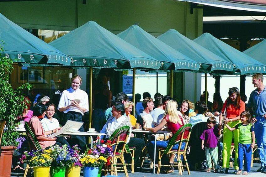 Un evento al aire libre con gente sentada en mesas bajo sombrillas verdes, rodeado de plantas y flores en macetas. 2