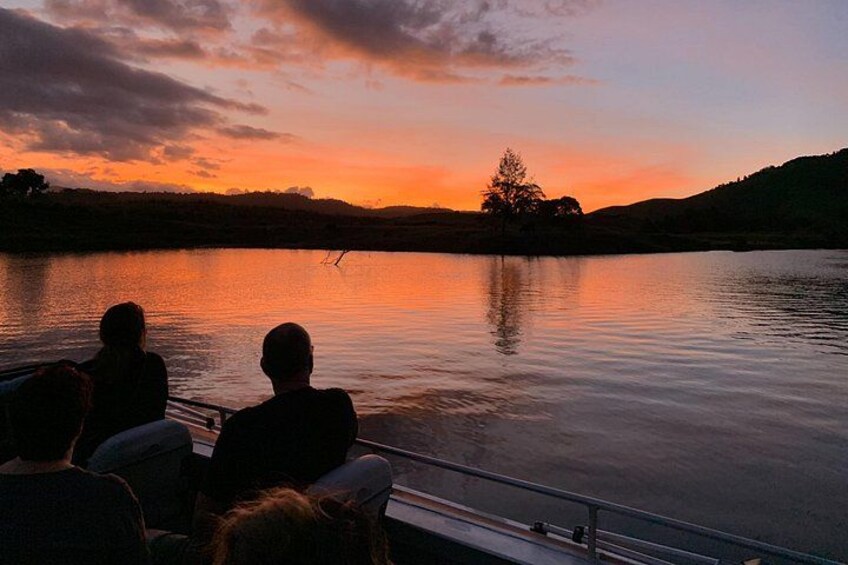 Ein Sonnenuntergang über einem ruhigen See mit Silhouetten von Menschen und einem Baum am Ufer. 2