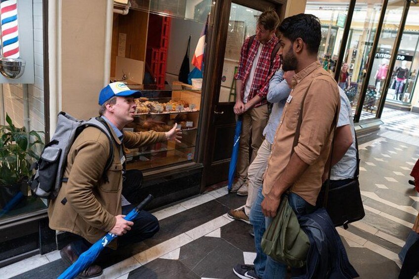 En person sidder på jorden foran en butik og holder en blå paraply, mens to andre personer står i nærheden. 1