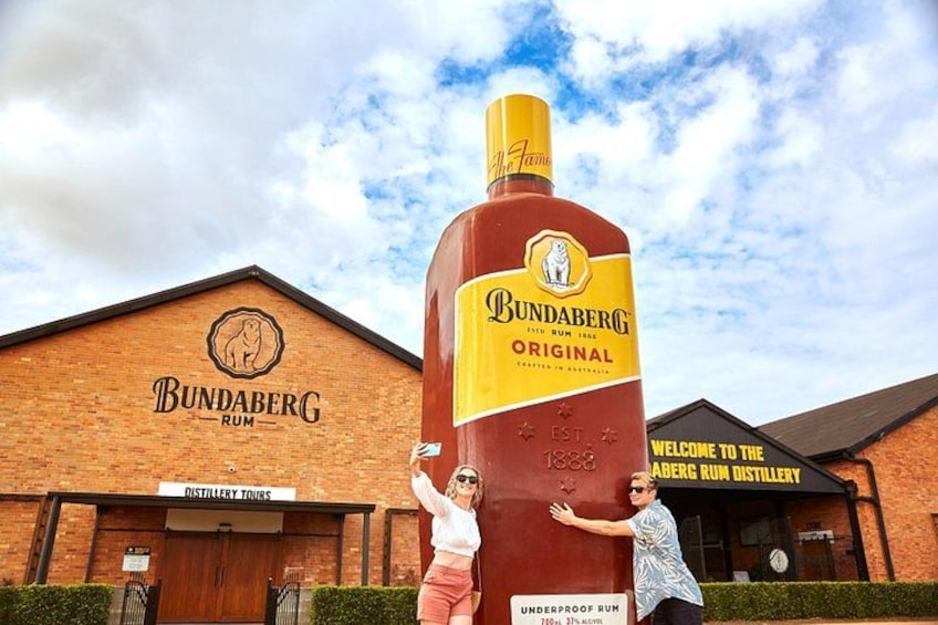 Une grande sculpture représentant une bouteille de rhum Bundaberg est placée devant un bâtiment en briques sur lequel est apposée une enseigne indiquant : « Visites guidées de la distillerie Bundaberg Rum ». 1