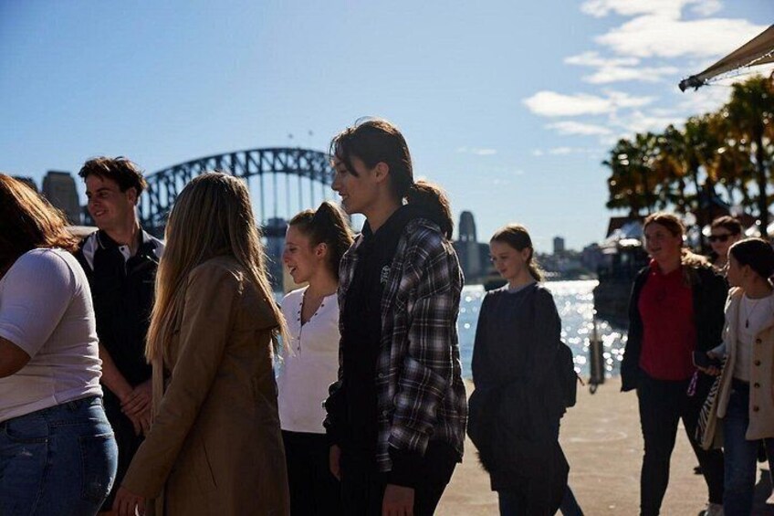 A group of people walking along a waterfront with a bridge in the background. 2