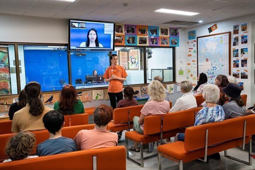A classroom with students seated on orange chairs, a teacher presenting at the front, and a large screen displaying a video. 5