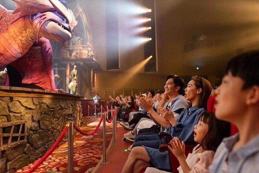 An audience seated in a theater watching a large animated dragon on stage. 1