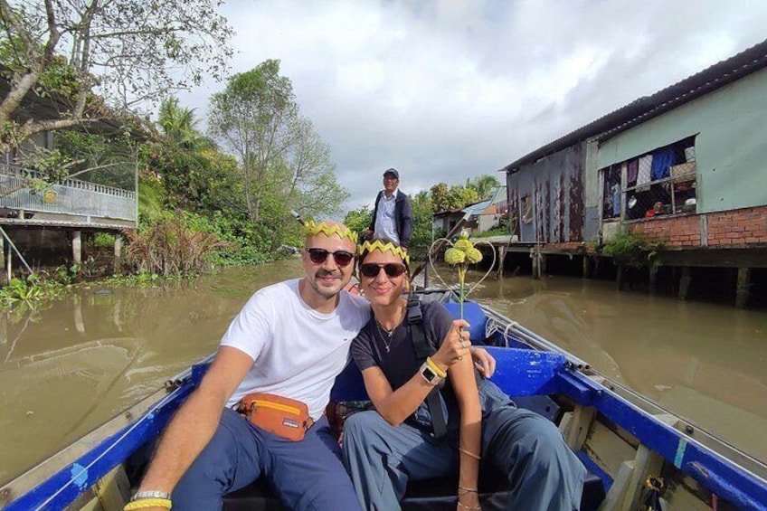 Two people in a boat on a river, surrounded by greenery and buildings. 1