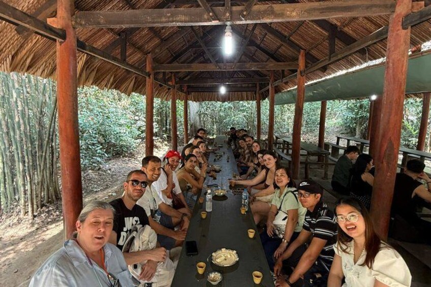 A group of people seated at a long table in an outdoor covered area with a thatched roof, surrounded by greenery. 3