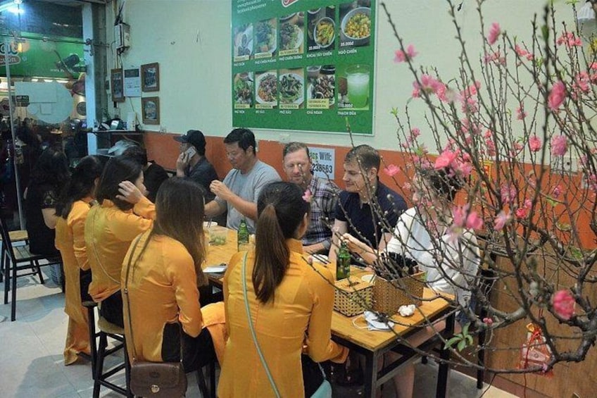 Un restaurante con comensales sentados en mesas, un tablero de menú en la pared y una planta decorativa con flores rosadas. 4