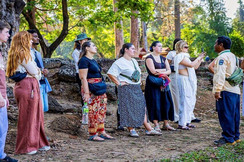 A group of people standing outdoors, some with backpacks, in a natural setting with trees and a dirt path. 3