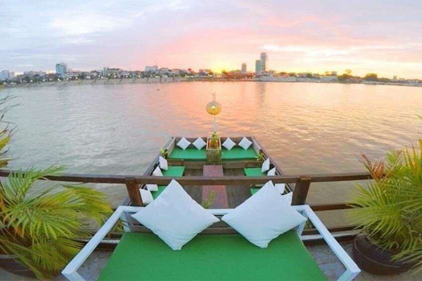 Un bote con cojines verdes y almohadas blancas, amarrado a orillas de un río con un horizonte urbano al fondo. 1