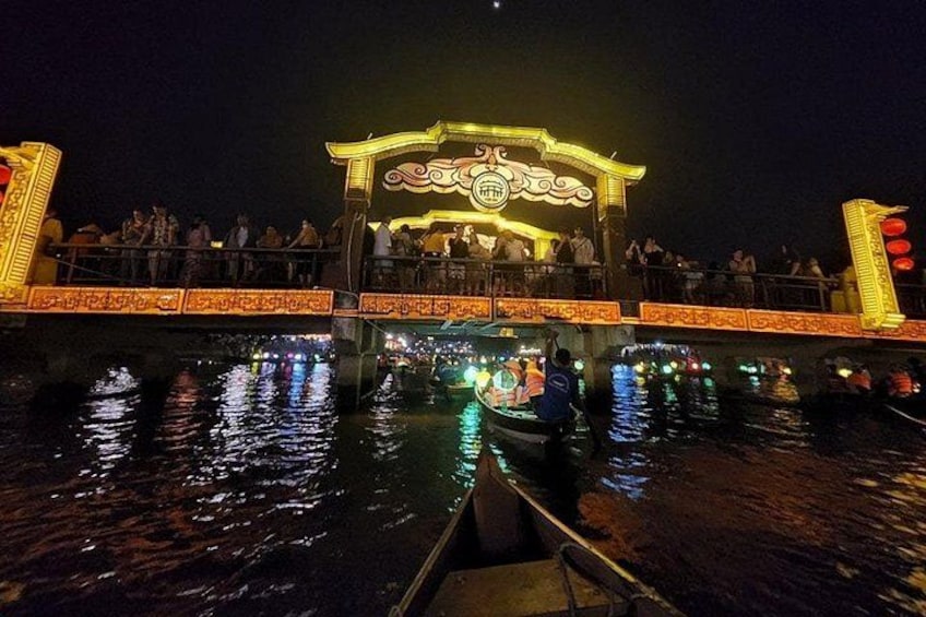 Ein traditionelles Boot mit beleuchteter Dekoration auf einem Fluss bei Nacht, eine Brücke mit aufwendigen Geländern und eine Menschenmenge auf der Brücke. 4