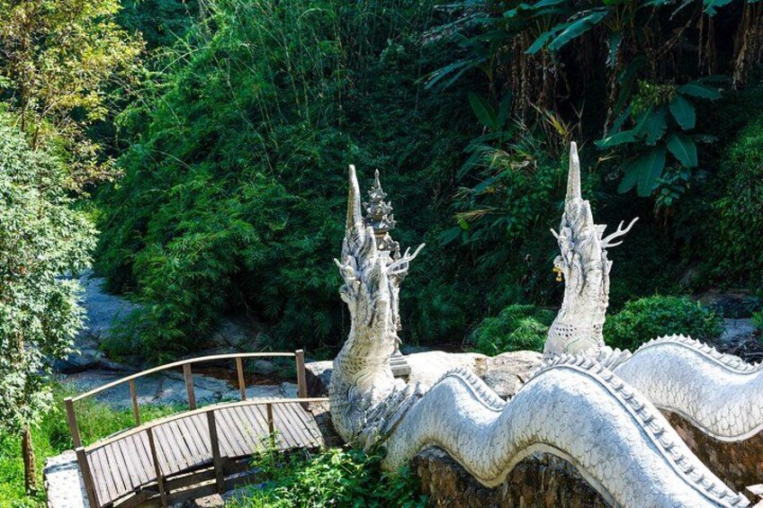Un puente de piedra con pasarela de madera, rodeado de abundante vegetación y dos estructuras de piedra ornamentales que recuerdan a criaturas míticas. 4