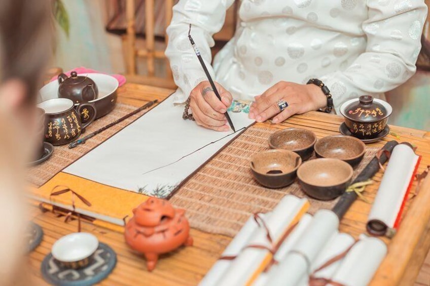 Una persona escribiendo en un papel con herramientas de caligrafía, rodeada de tradicionales conjuntos y cuencos de té chinos. 1