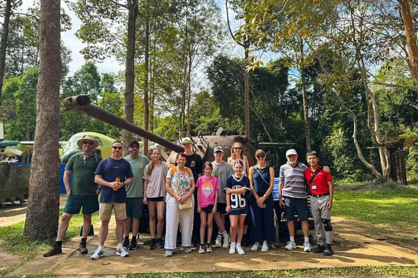 A group of people standing outdoors with trees and a large structure in the background. 3